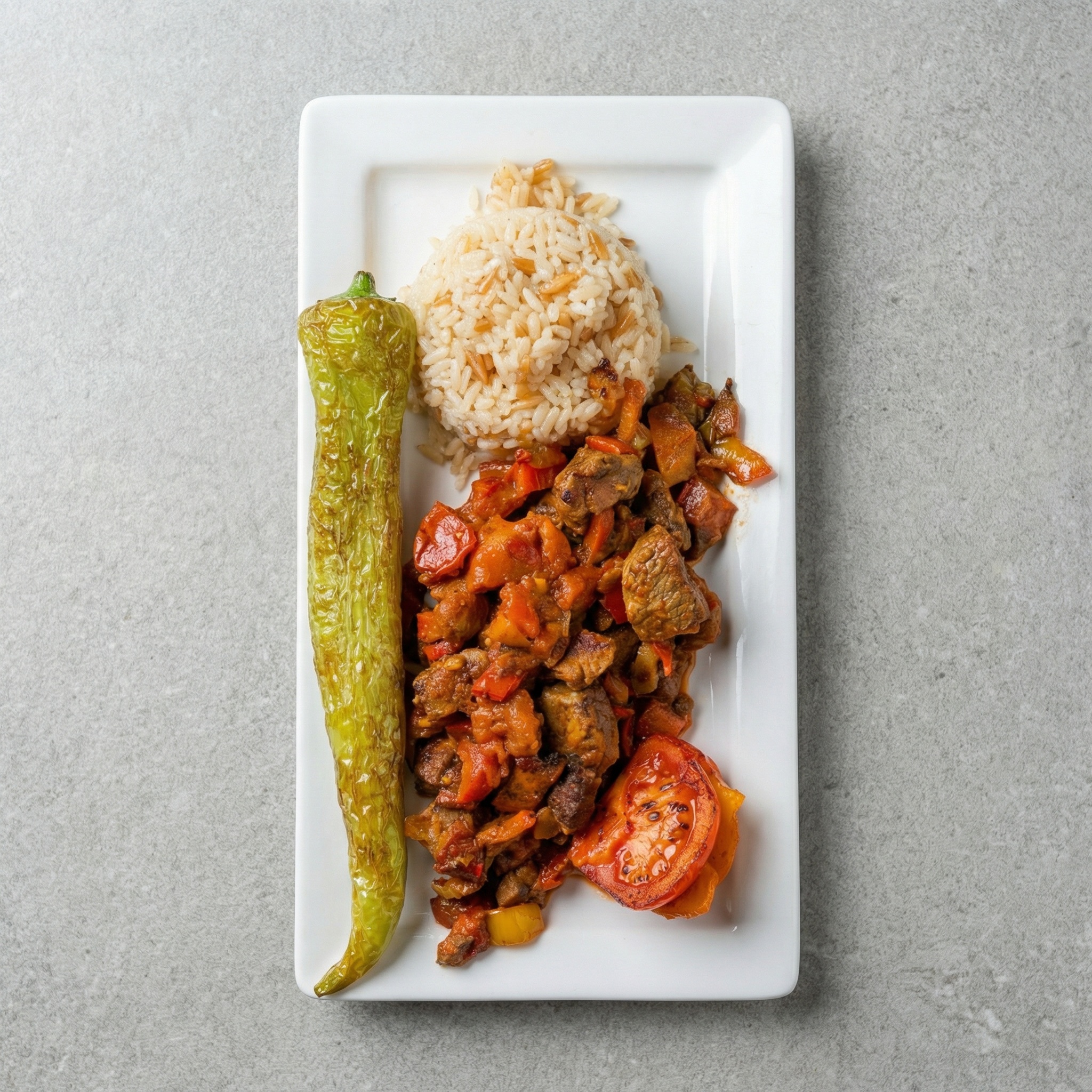 Ein rechteckiger weißer Teller mit einer Portion Reis, würzigem Fleisch-Gemüse-Sote, einer Grillpaprika und einer gegrillten Tomate auf grauem Hintergrund.