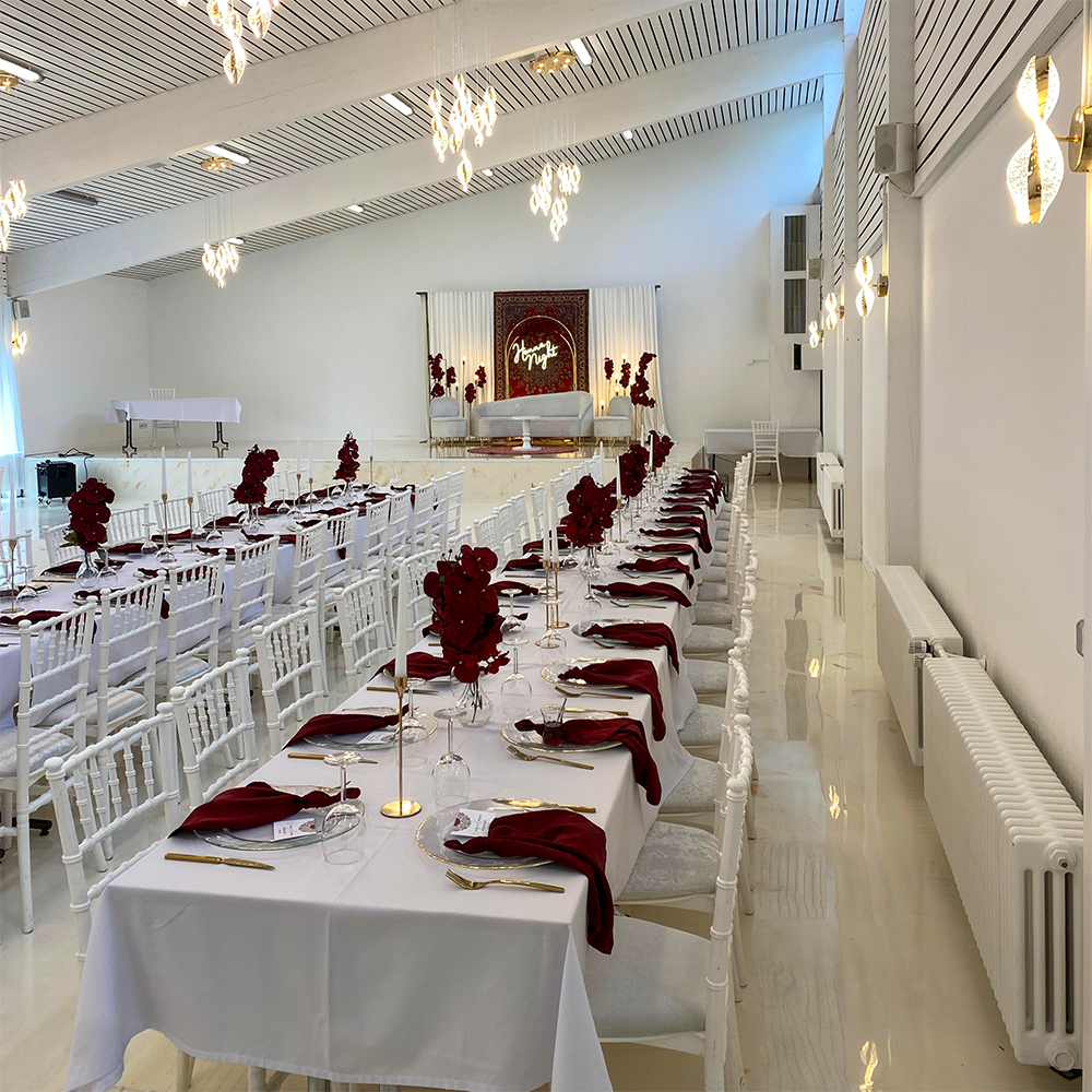 Festlich dekorierter weißer Festsaal mit langen Tafeln und roten Blumen.