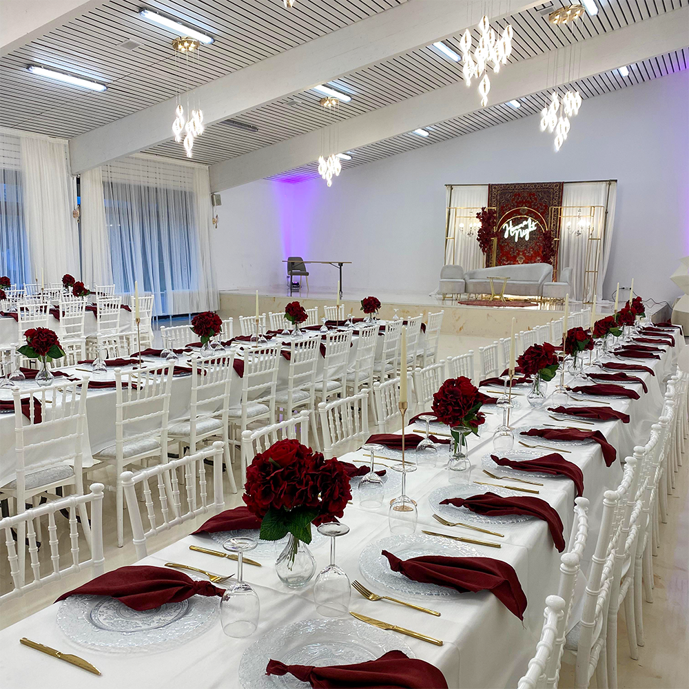 Festlich dekorierter weißer Festsaal mit langen Tafeln und roten Blumen.